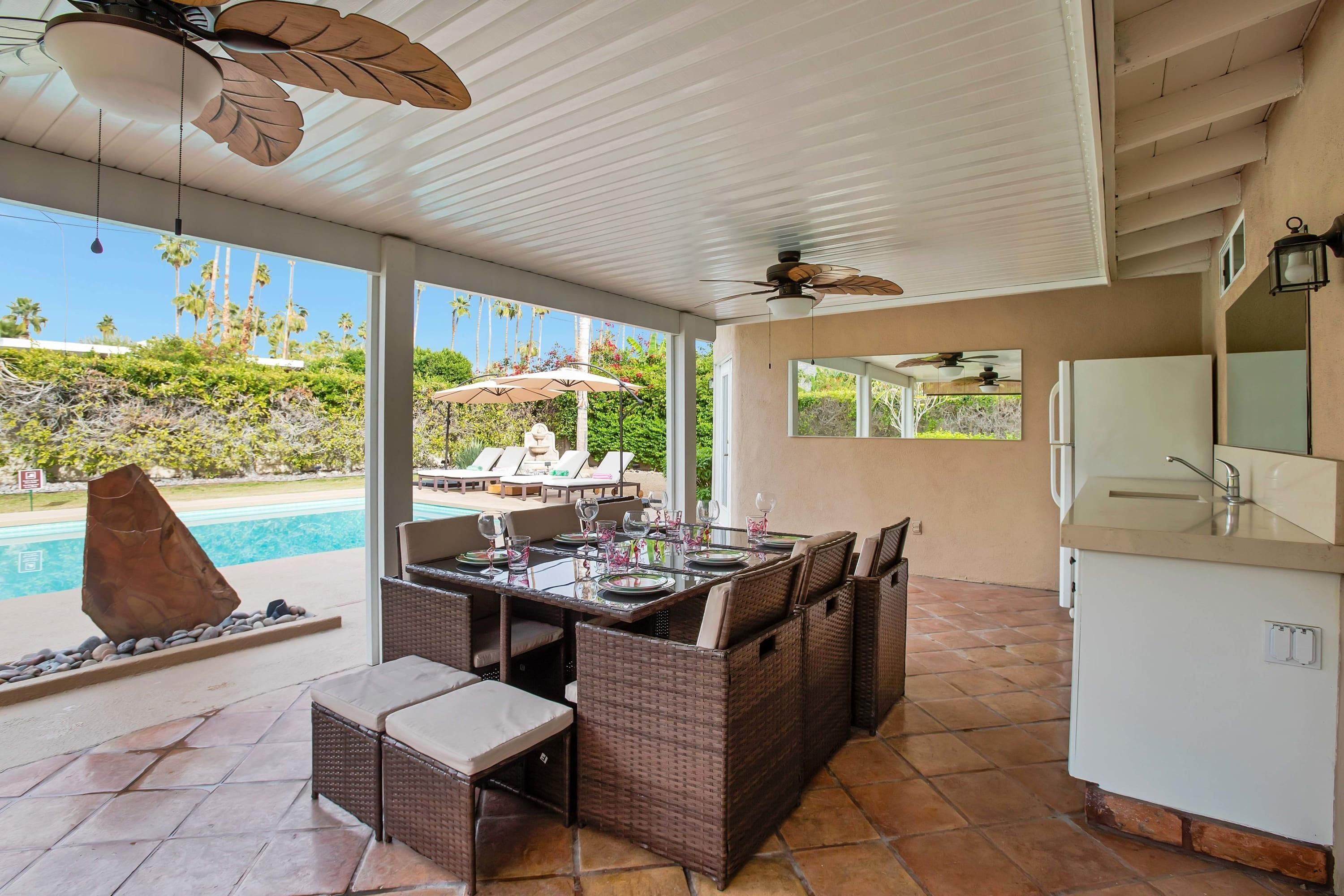 A covered outdoor dining area with wicker furniture, a pool, and a kitchen area with a refrigerator and sink.