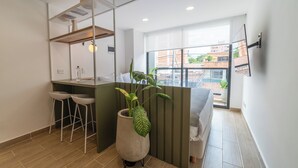 Une pièce moderne et bien éclairée, avec un plancher en bois, un bureau agrémenté de deux chaises blanches, une grande fenêtre donnant sur des immeubles et une plante en pot.