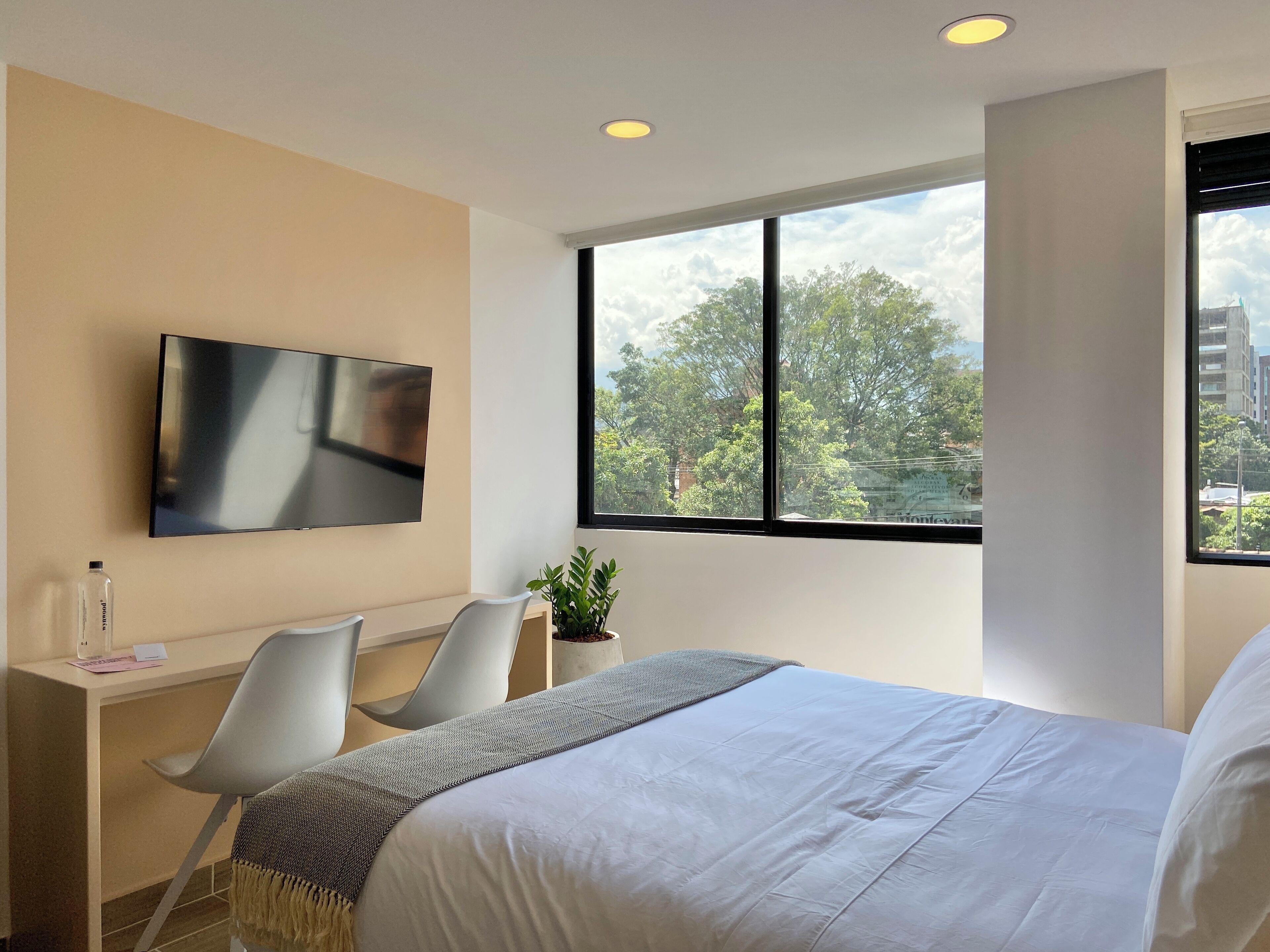 Una habitación de hotel moderna con una cama, un escritorio con silla, un televisor y un ventanal con vistas a árboles y edificios.