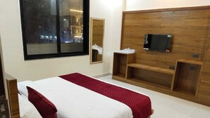 Une chambre moderne avec une tête de lit en bois, un lit avec une literie blanche et rouge, un téléviseur fixé sur la tête de lit et une fenêtre donnant sur la ville.