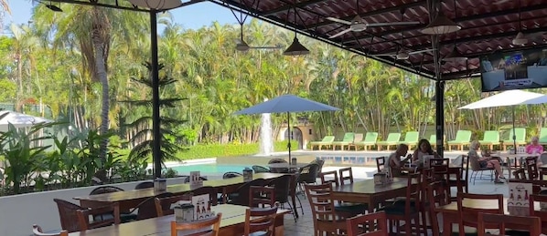 An outdoor dining area with tables and chairs, a pool, and palm trees in the background. - The Palms At Coco Beach (Carrillos)