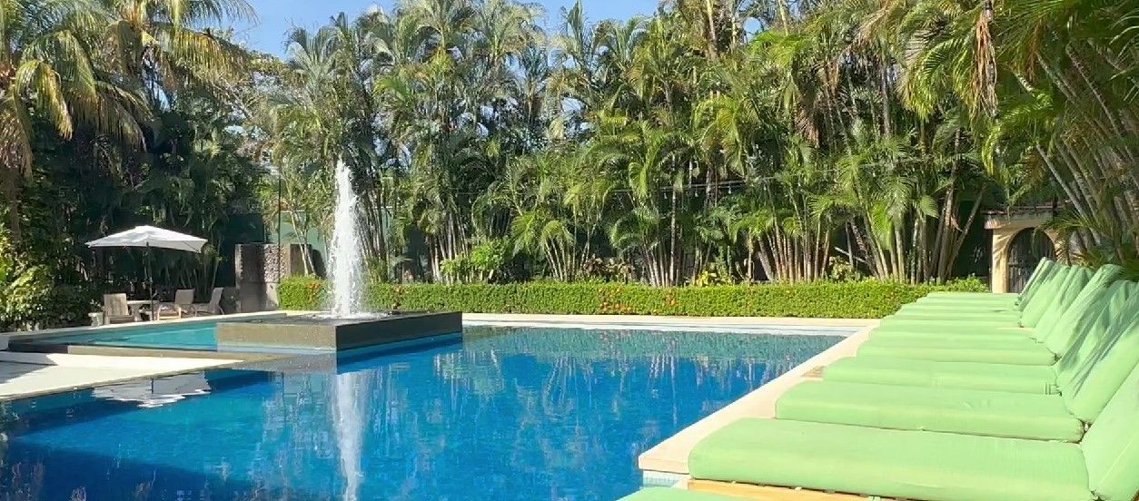 Una piscina con una fuente, rodeada de abundante vegetación y una fila de sillas de descanso verdes.