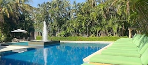 Una piscina con una fuente, rodeada de abundante vegetación y una fila de sillas de descanso verdes.
