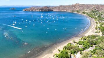 Vista aérea de una zona costera con playa, barcos y montañas al fondo.