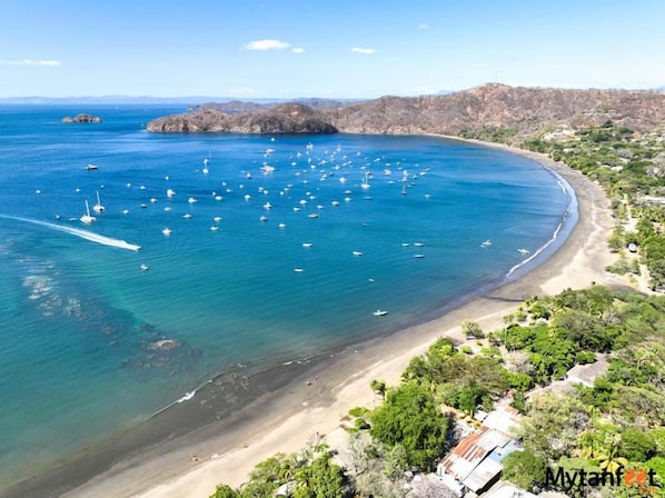 Vista aérea de una zona costera con playa, barcos y montañas al fondo.