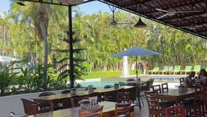 An outdoor dining area with tables and chairs, a pool, and palm trees in the background. - The Palms At Coco Beach (Carrillos)