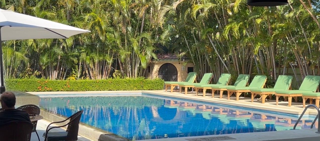 Una piscina con sillones de descanso, rodeada de palmeras y un edificio con tejado de baldosas.