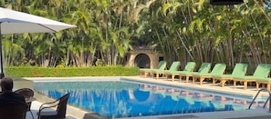 Una piscina con sillones de descanso, rodeada de palmeras y un edificio con tejado de baldosas.