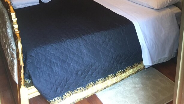 Ein ordentlich bezogenes Bett mit einer dunkelgrauen Steppdecke, weißen Bettwäsche und einem goldverzierten Kopfteil, darüber ein gerahmtes Bild.