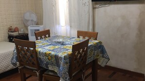 Ein Essbereich mit Tisch, der mit einer gemusterten Tischdecke bedeckt ist, umgeben von Holzstühlen. Eine weiße Klimaanlage befindet sich an der Wand, und ein Fernseher ist über dem Tisch an der Wand montiert.
