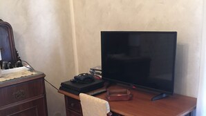 Ein Holztisch mit einem Fernseher, einer Fernbedienung und einem Spielcontroller. Dazu gehören ein Stuhl mit Kissen, eine Kommode und eine Lampe.