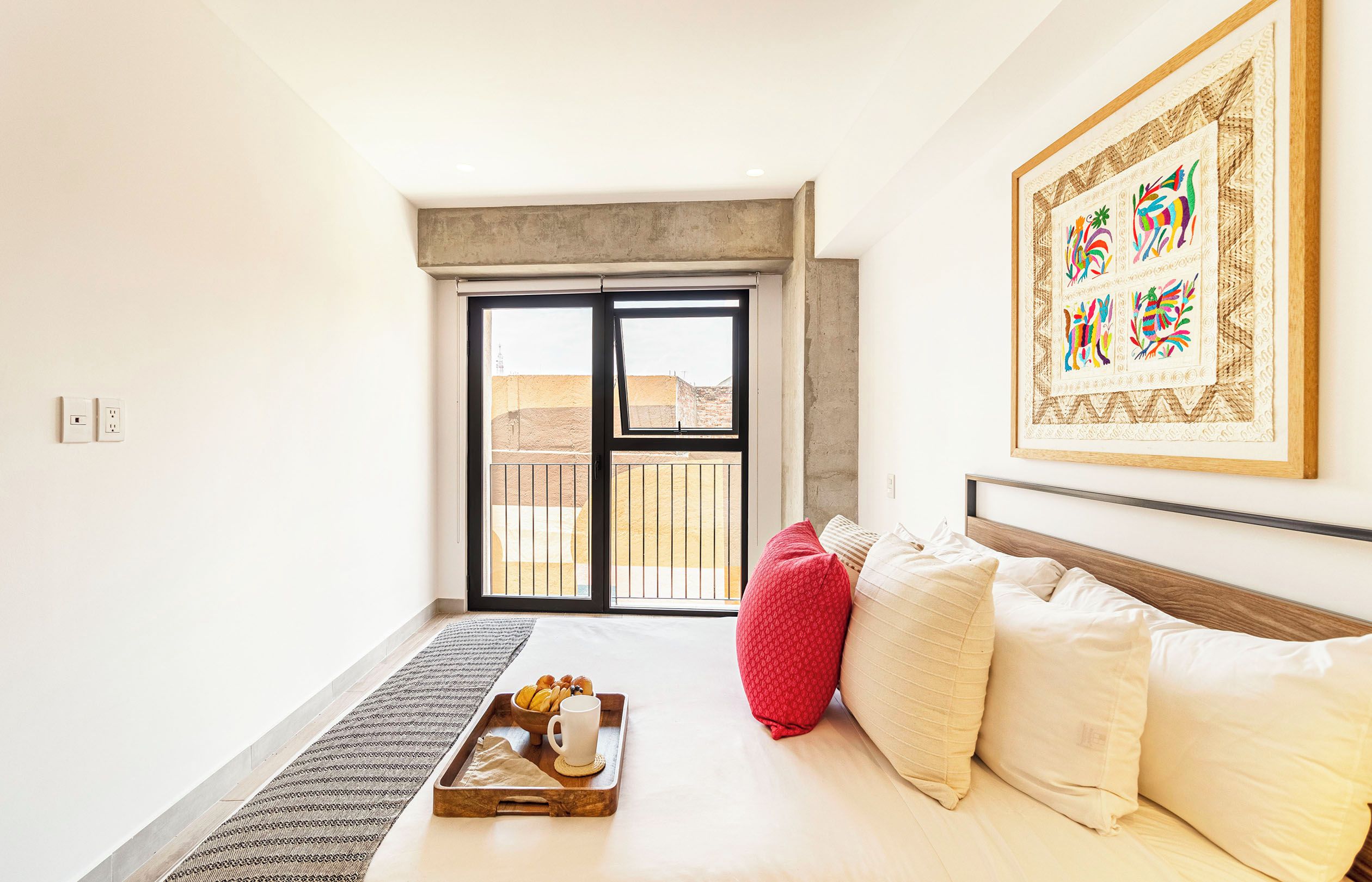 Habitación de hotel con cama, sillón de peluche rojo, bandeja con pasteles y una taza, una obra de arte grande enmarcada y vista por una puerta corredera.