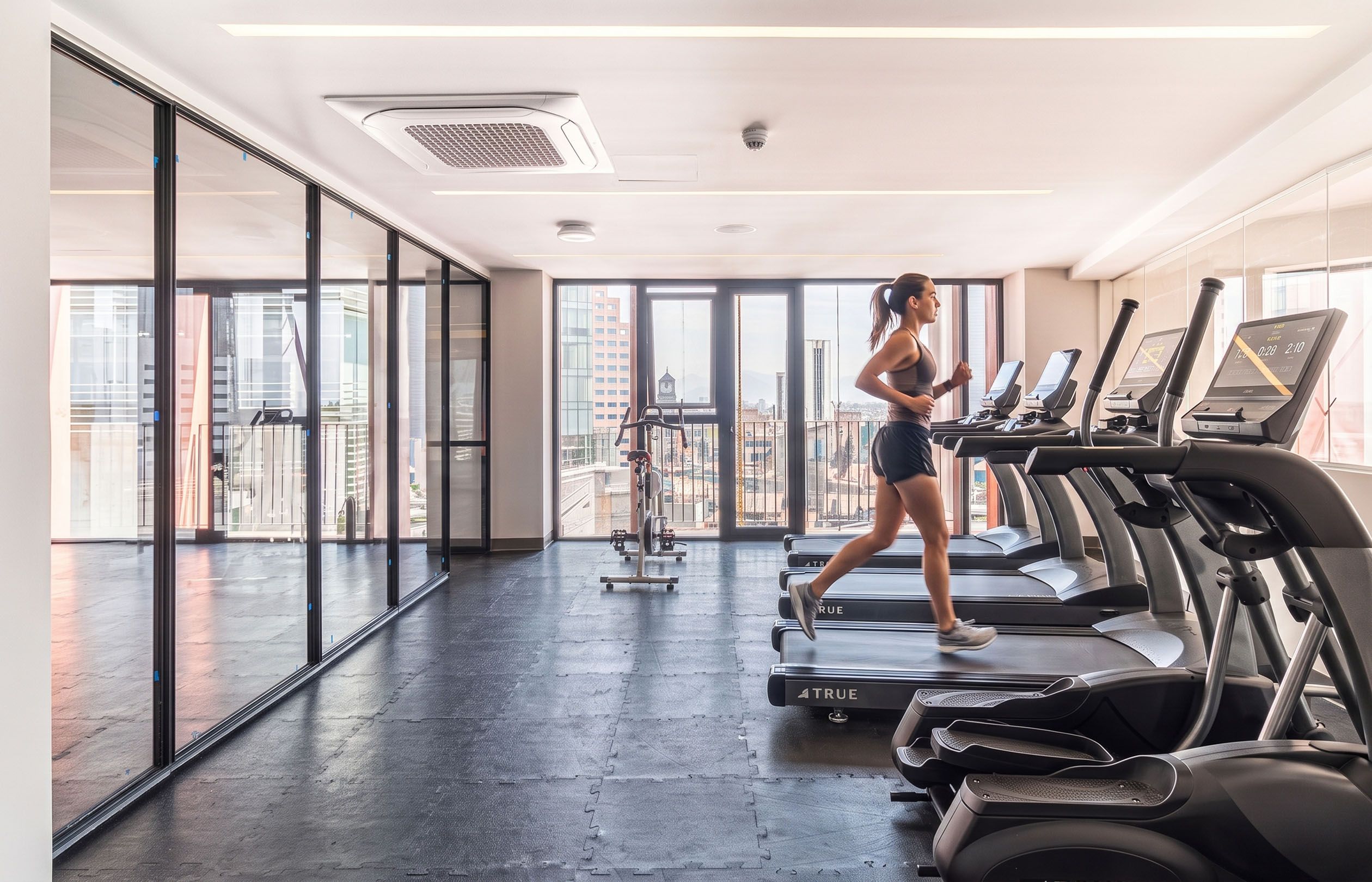 Un gimnasio con cintas de correr, una máquina de remo y grandes ventanales con vista a la ciudad.