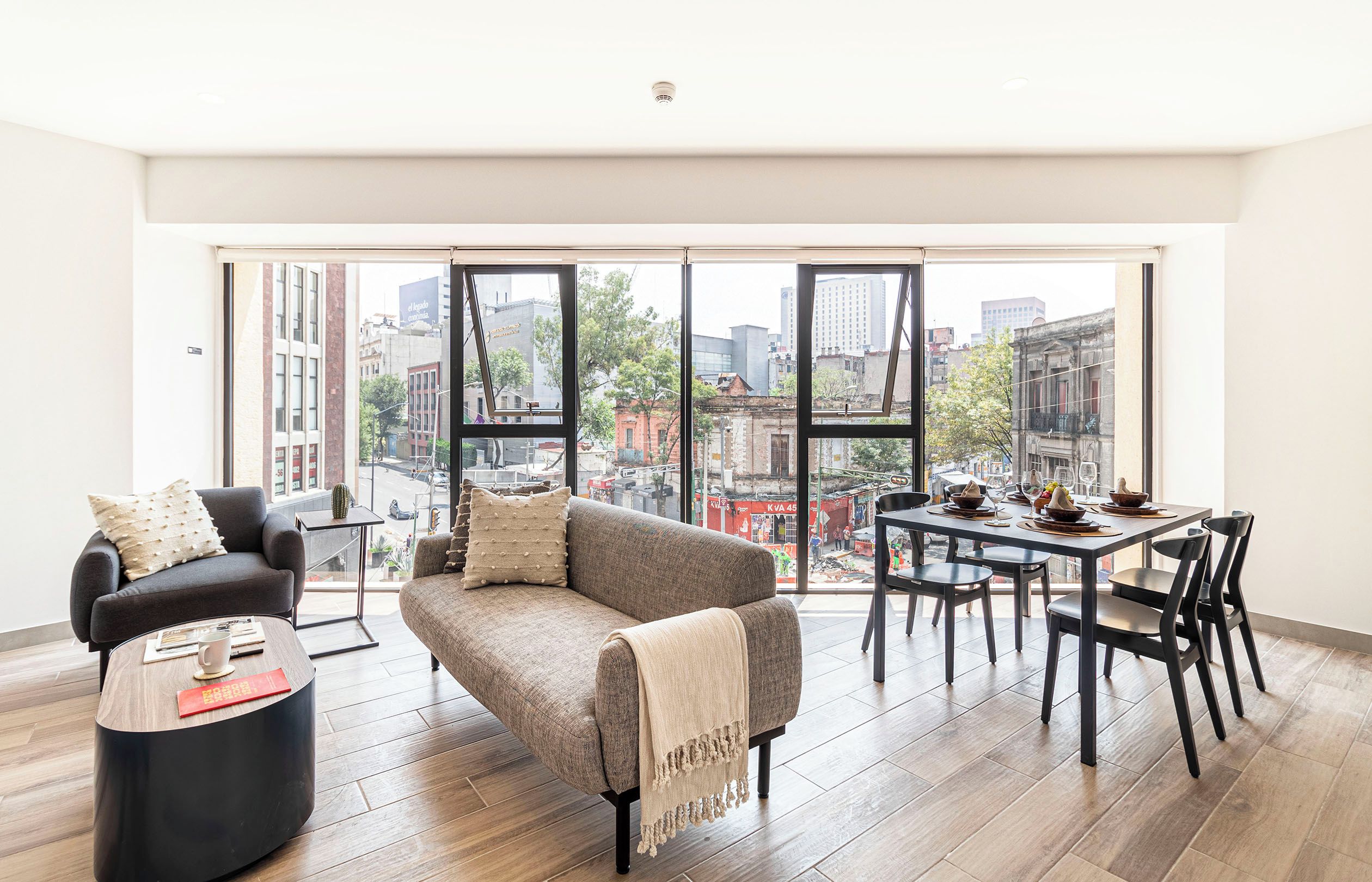Amplia sala de estar con sofá, mesa de centro y comedor con sillas y una mesa dispuesta para una comida. Amplias ventanas ofrecen vista a la ciudad.