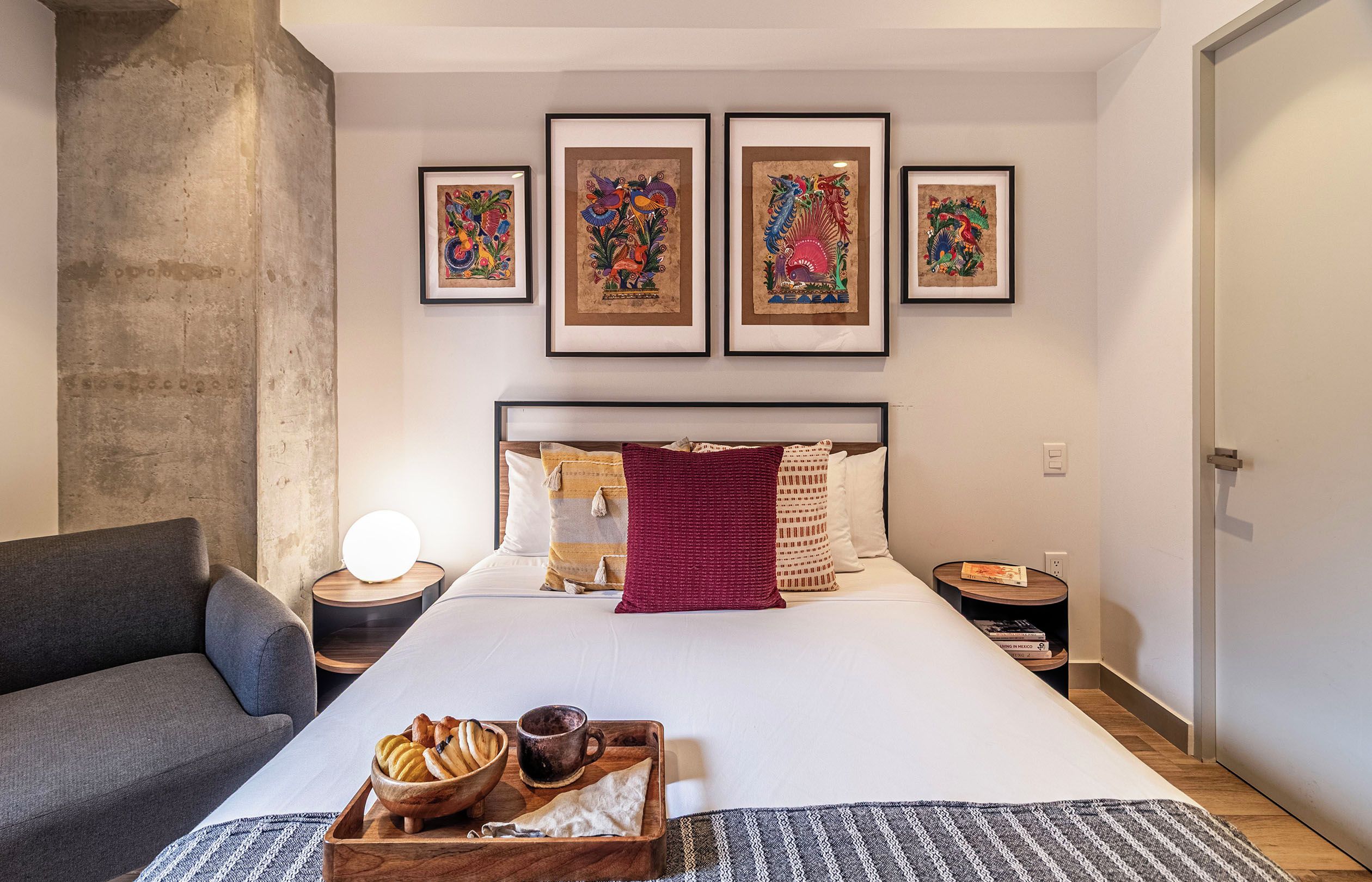 Une chambre d’hôtel moderne avec un grand lit, une table de chevet avec une lampe, une petite table d’appoint avec des livres et un mur orné de quatre œuvres d’art encadrées.