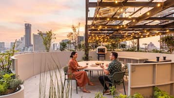 Un área de comedor en la azotea con vistas a la ciudad, mesas al aire libre y luces colgantes.