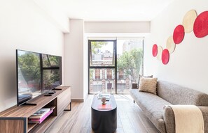 Una sala de estar moderna con un televisor de pantalla plana, una mesa consola de madera, una mesa redonda de centro, un sofá gris y vistas de un paisaje urbano a través de una puerta de cristal.