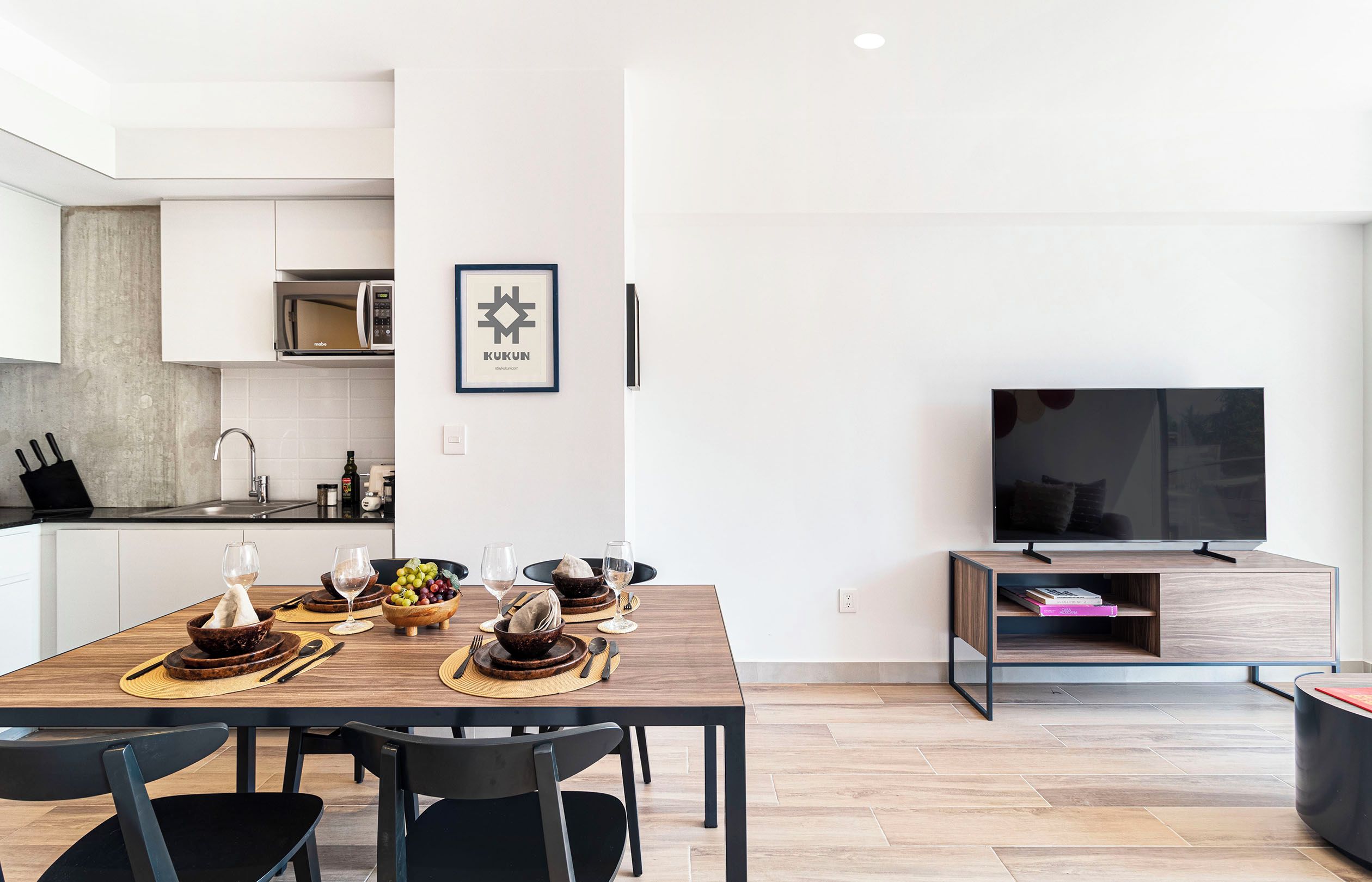 Una cocina moderna con microondas, mesa de comedor dispuesta con platos, tazones y vasos, y un televisor de pantalla plana sobre un pedestal.