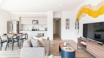 Una sala de estar moderna con un sofá, una mesa de centro y un televisor. Se ve una cocina al fondo con gabinetes y electrodomésticos blancos.