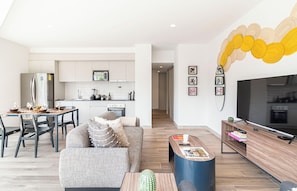 A modern living room with a sofa, coffee table, and a TV. A kitchen with white cabinets and appliances is visible in the background. - Kukun Reforma Downtown (Mexico City)