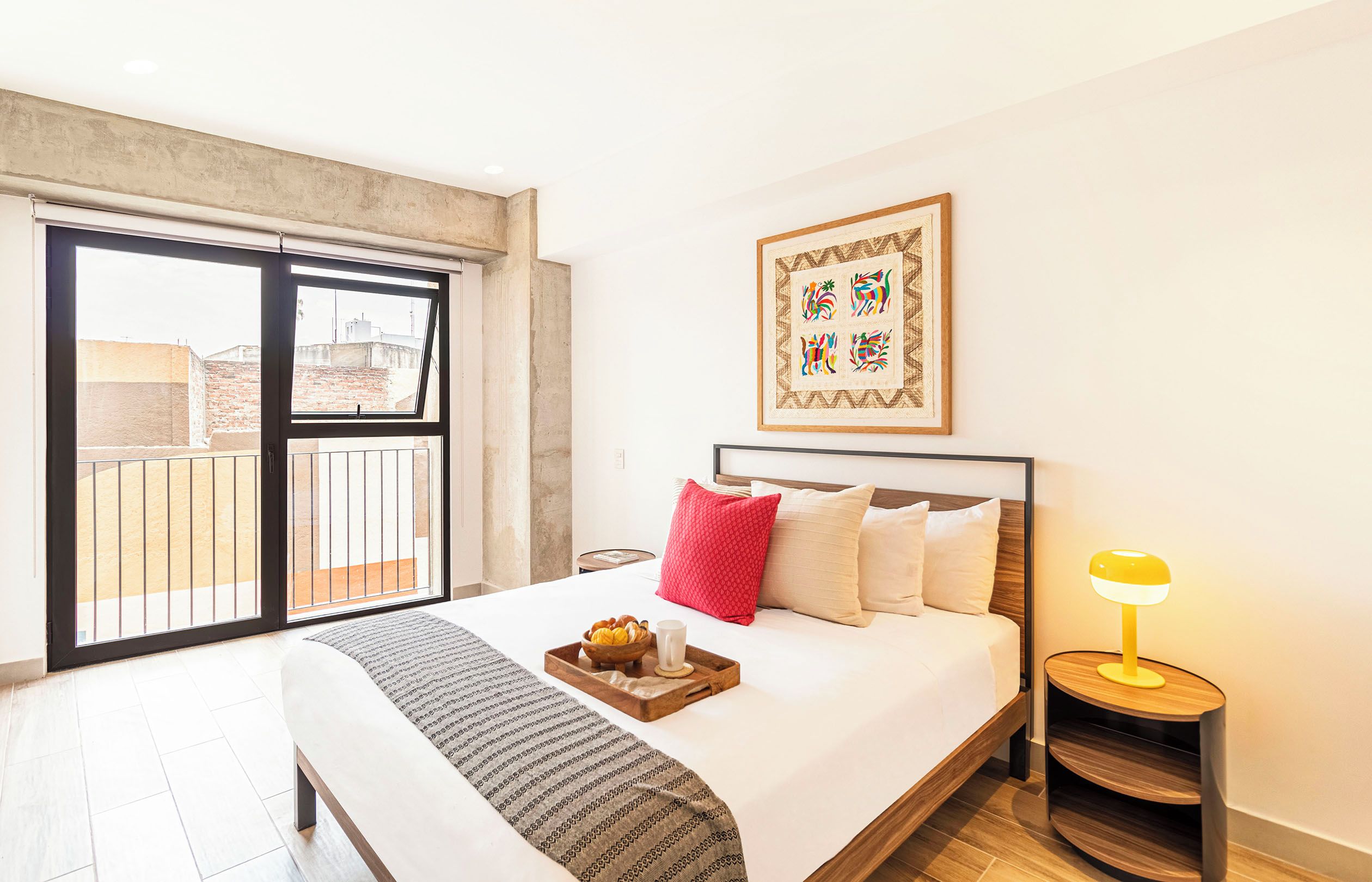 Quarto de hotel com uma cama, mesa de cabeceira com abajur, janela com vista para edifícios e quadro na parede.