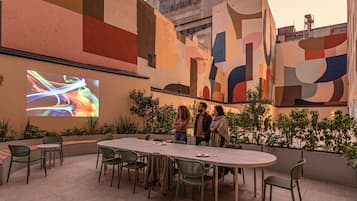 Un área de comedor al aire libre con una mesa grande y sillas, rodeada de follaje y un mural colorido en la pared.
