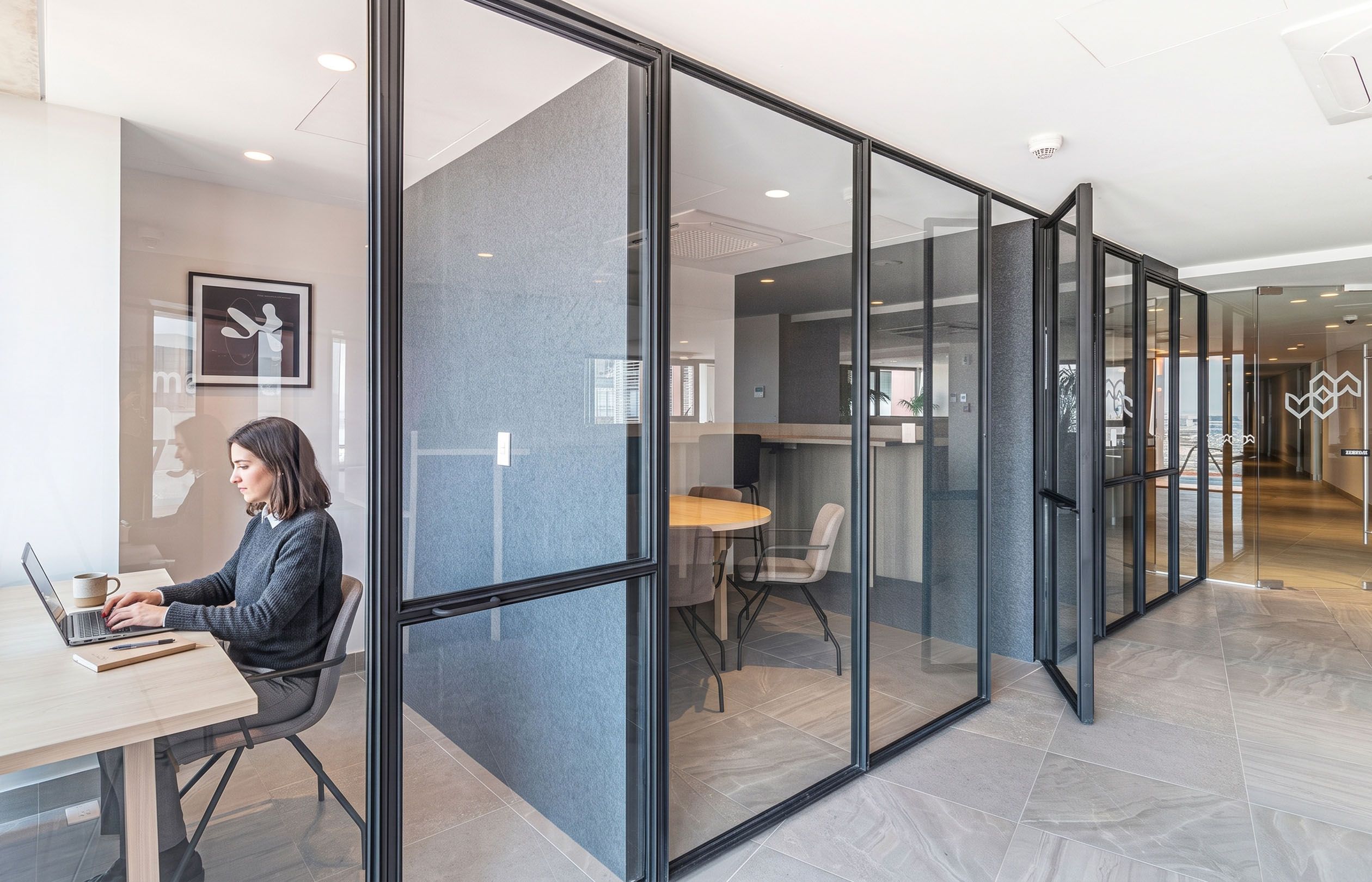 Un espace de bureau avec des cloisons en verre, une personne travaillant à un bureau et une vue sur le paysage urbain à travers le verre.