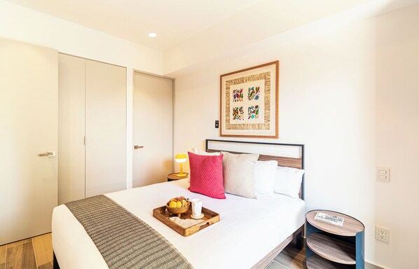 Una habitación de hotel con cama, una mesita de noche con lámpara, una mesita redonda pequeña y una obra de arte colgada en la pared.
