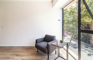 A modern room with a grey sofa, a small wooden table, and a large window offering a view of the street. - Kukun Reforma Downtown (Mexico City)
