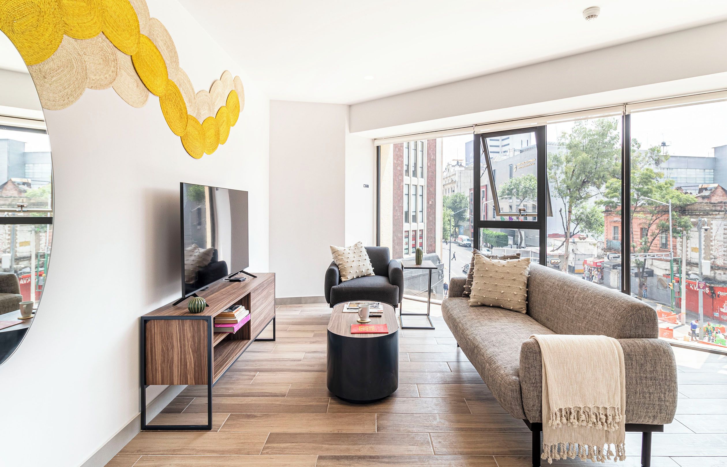 Una sala de estar moderna con sofá, mesa de centro, televisión y vista a la ciudad a través de amplios ventanales.