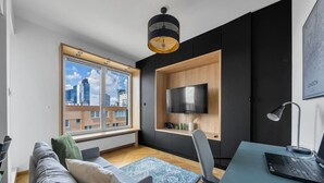 A modern living room with a large window, a flat-screen TV mounted on a wooden shelf, a desk with a laptop, and a sofa with cushions. - Warsaw Wola Studio - Balcony & Workspace -by Rentujemy (Warszawa)