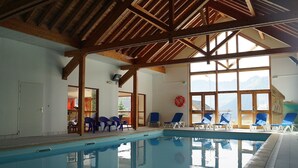 Piscina interior com vigas de madeira, espreguiçadeiras azuis e grandes janelas com vista para as montanhas.
