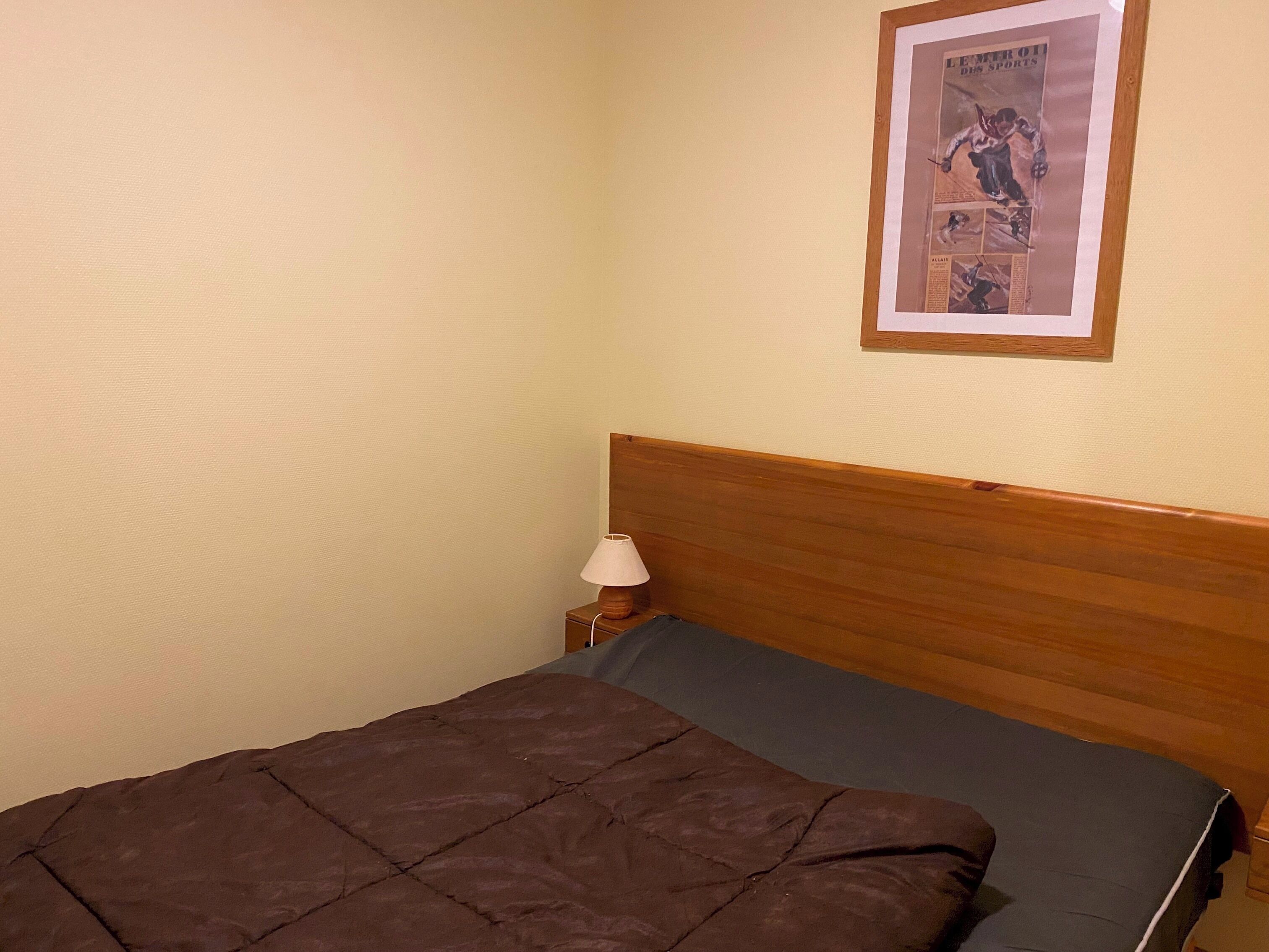 Quarto com cama, cabeceira de madeira, mesa de cabeceira com luminária e pôster esportivo emoldurado na parede.