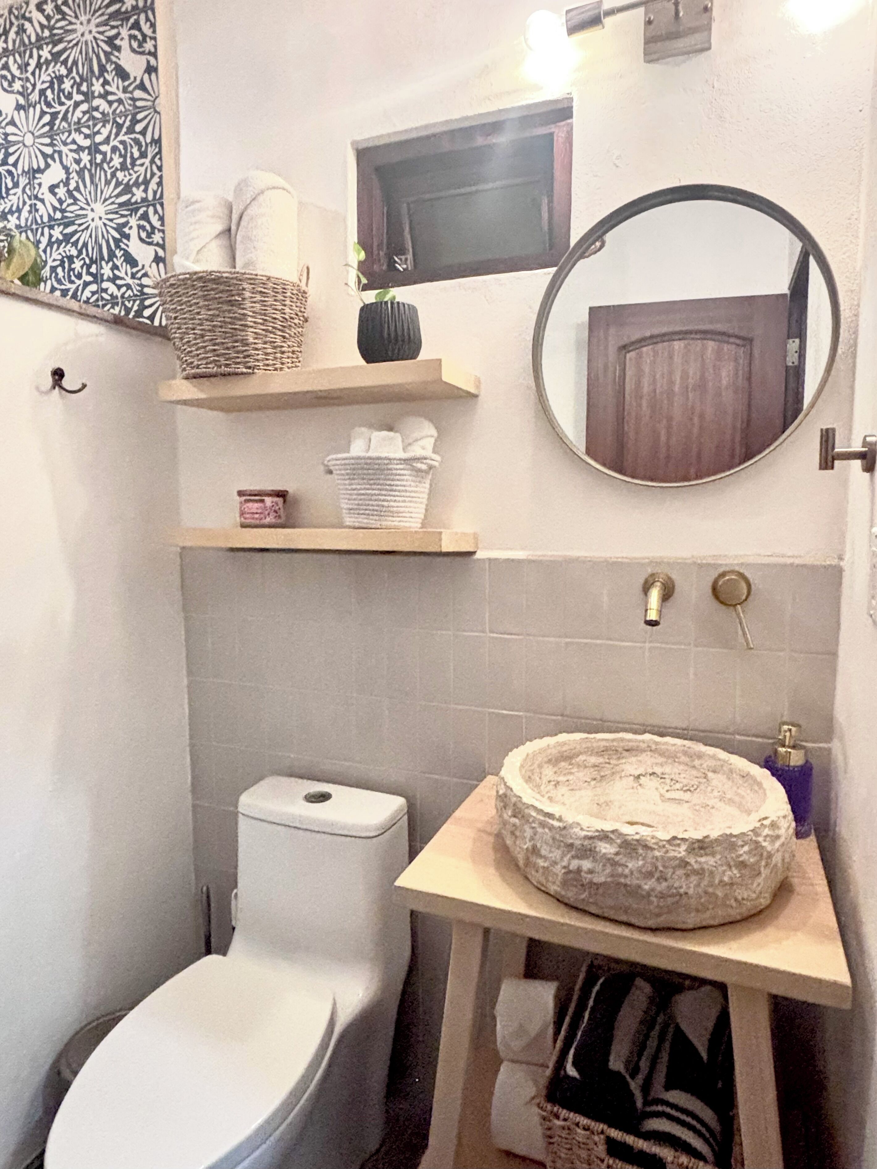 Une salle de bain comprenant des toilettes, un lavabo avec un receveur en pierre, un miroir et des étagères avec des serviettes et des paniers.