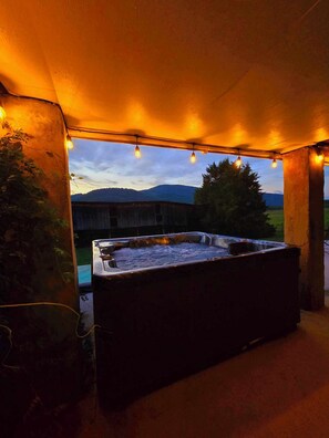 Un jacuzzi bajo una zona techada con luces colgantes, rodeado de un paisaje natural y una estructura de madera al fondo.