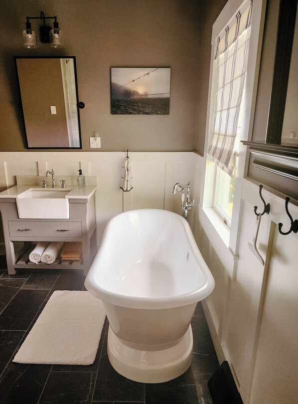 Une salle de bain avec une baignoire sur pieds, un meuble-lavabo avec un miroir et une fenêtre avec des stores.