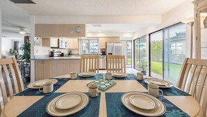 A modern kitchen with wooden cabinets, a dining area with a table set for four, and large windows offering a view of the outdoors. - Waterfront Villa: Gulf Access Canal,  Dog Friendly, Heated Pool (Cape Coral)