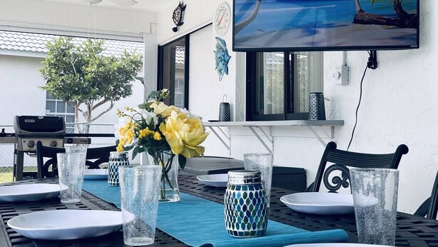 A dining area with a table set for two, featuring clear glasses, a vase with yellow flowers, and a blue table runner. - Waterfront Villa: Gulf Access Canal,  Dog Friendly, Heated Pool (Cape Coral)