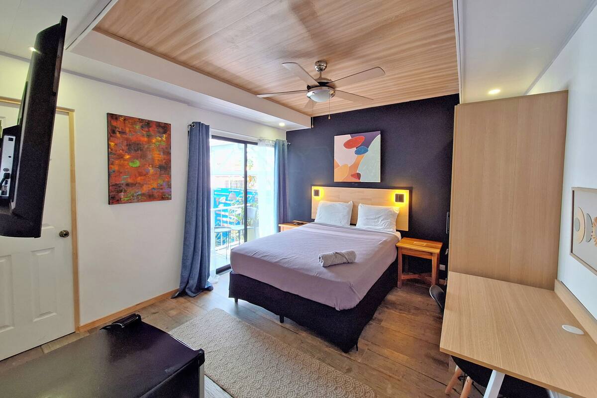 Un dormitorio moderno con cama, mesitas de noche, escritorio, ventilador de techo y obras de arte en las paredes.