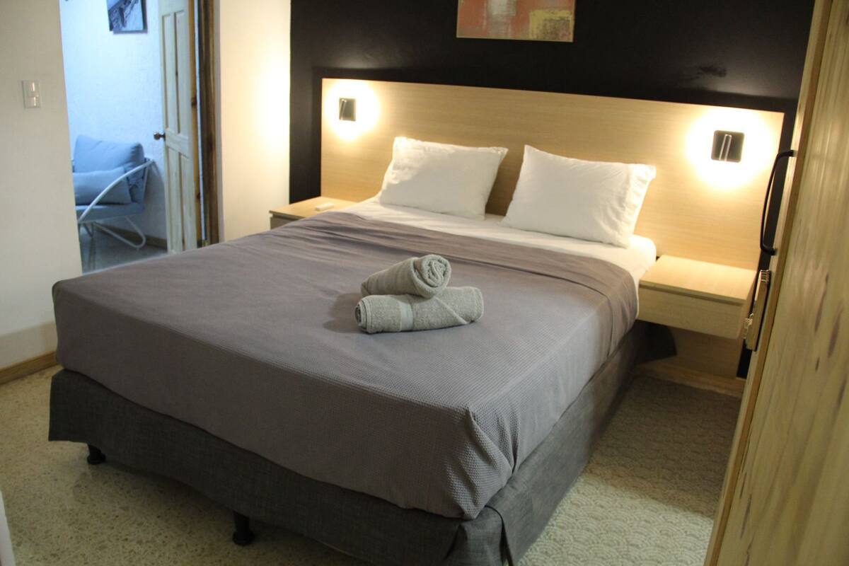 Una habitación de hotel con cama, mesita de noche, lámpara de pared y puerta que da a otra habitación.