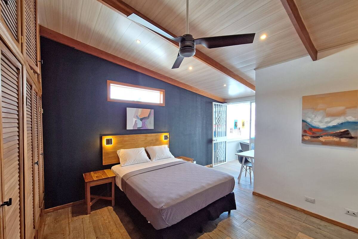 Un dormitorio con cama, mesita de noche, ventilador de techo, obras de arte y vistas al exterior.