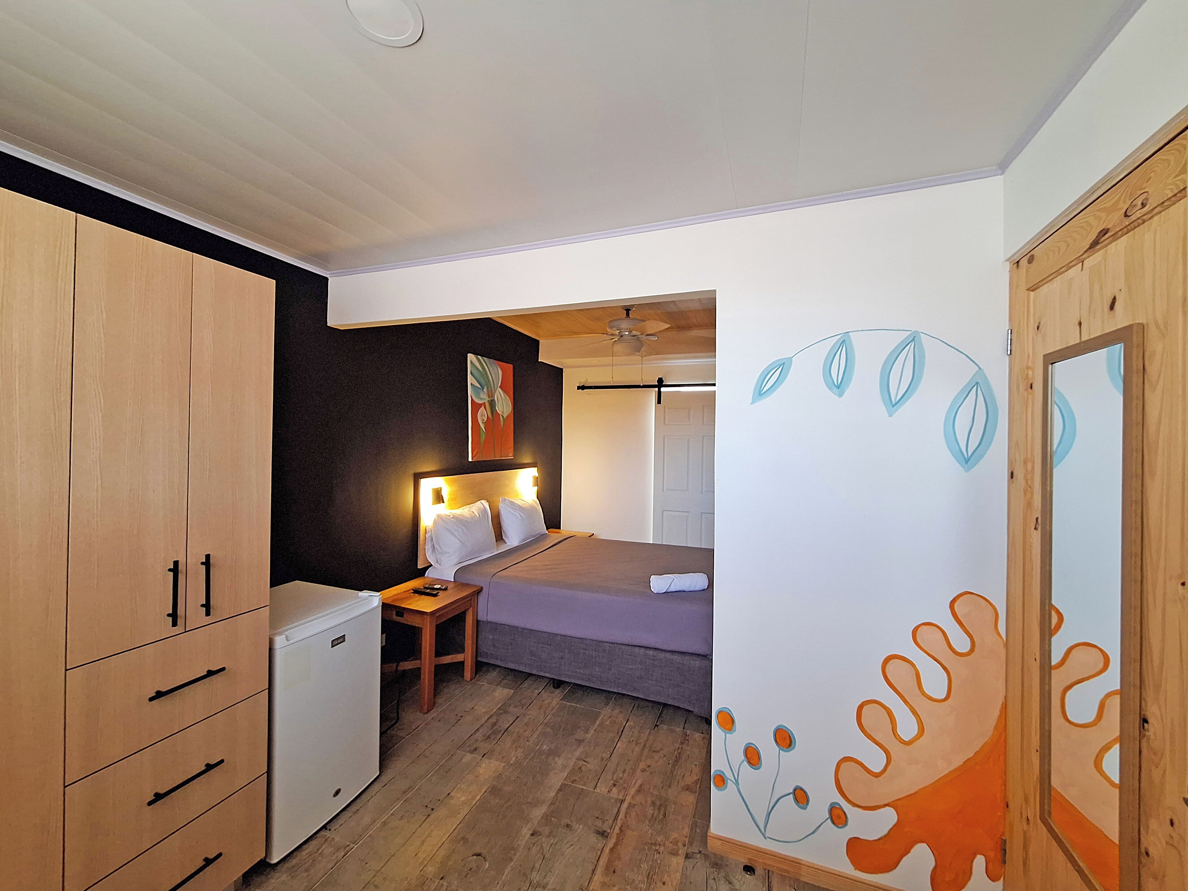 A compact bedroom with a bed, bedside table, and a small desk with a chair. There is a wardrobe with multiple drawers and a ceiling fan.