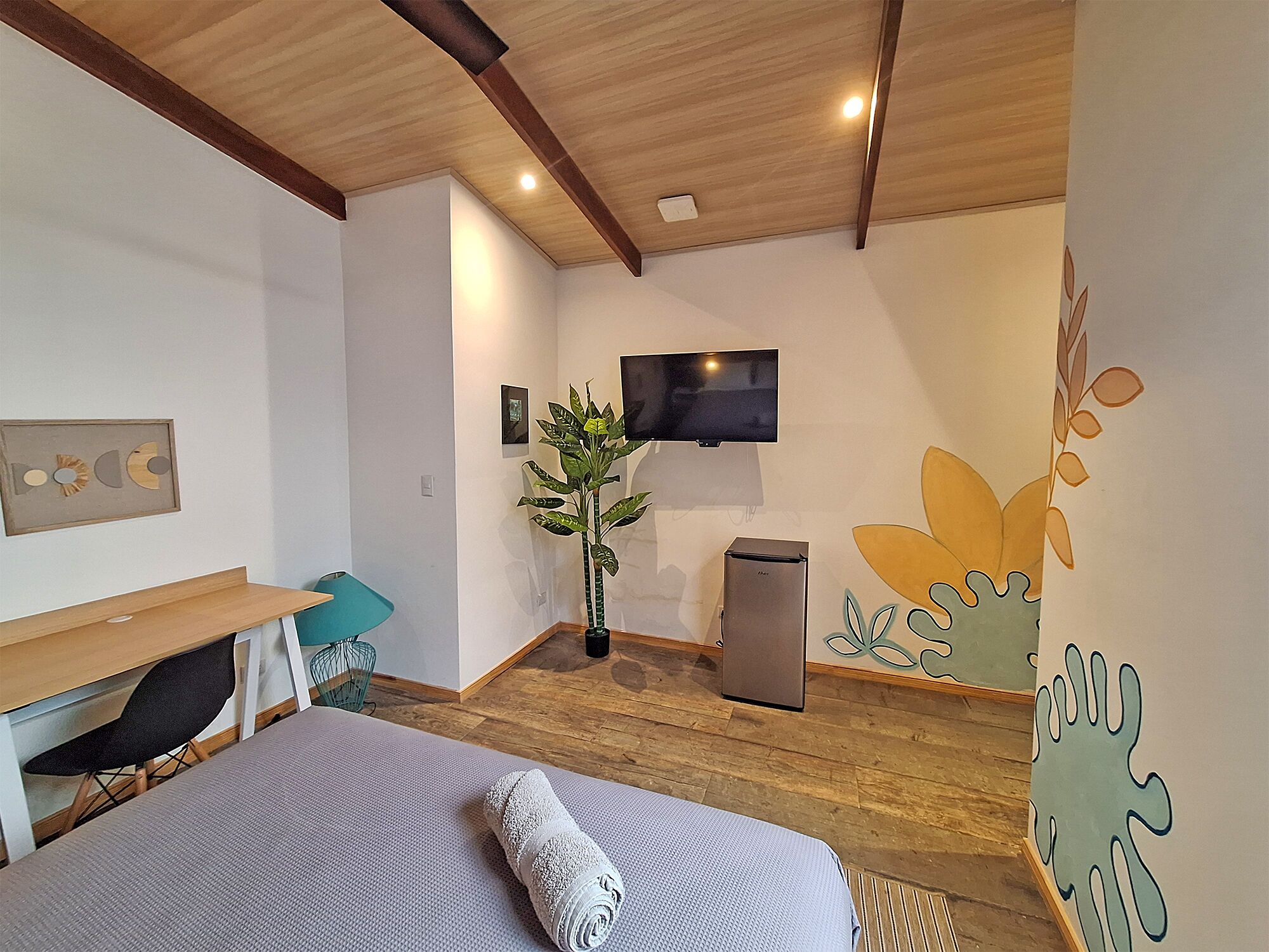 Habitació d'hotel moderna amb sostre de fusta, un llit amb una tovallola enrotllada, un escriptori amb una cadira, una televisió i una paret amb un dibuix de fulles.