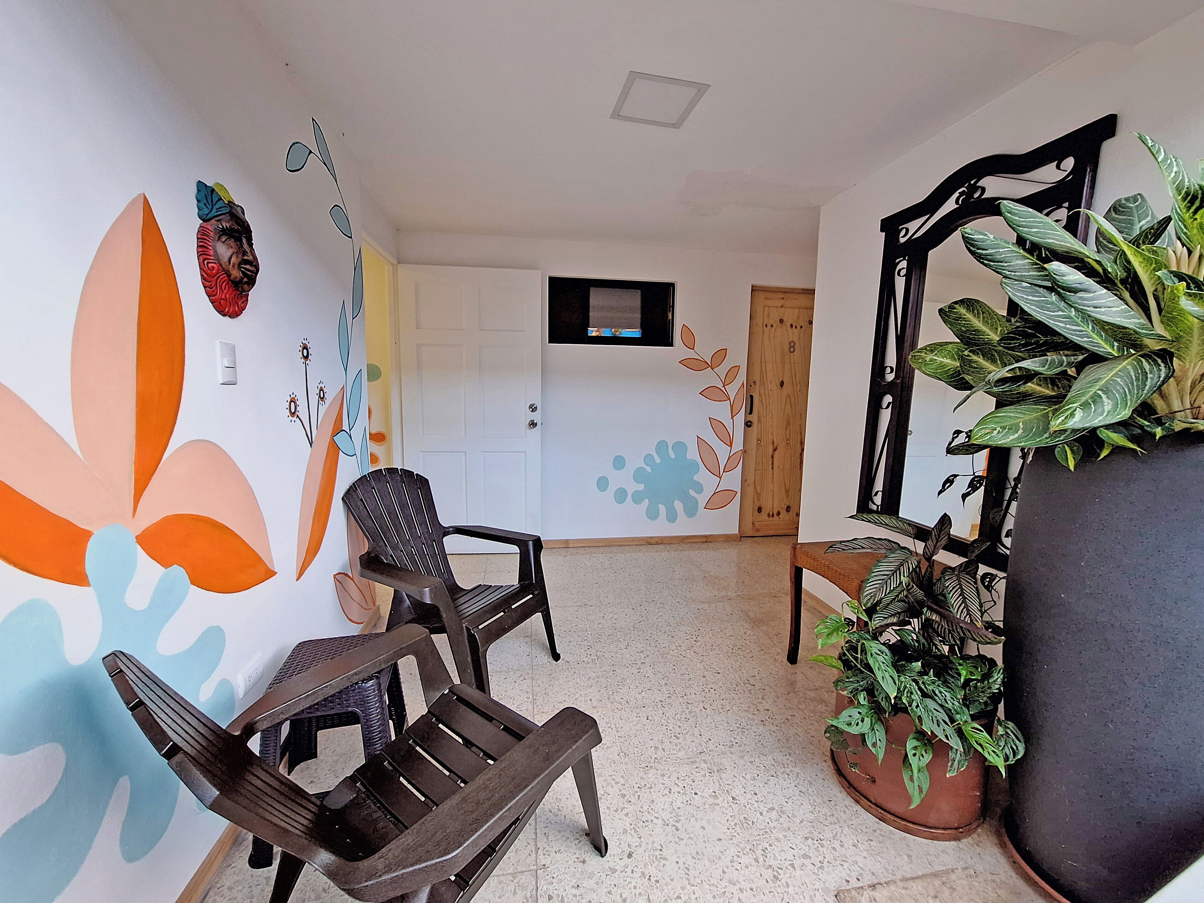 A room with a large potted plant, a wooden chair, a wall with a floral mural, and a door with a number.