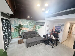 A living area with a sofa, ottoman, and a desk with a chair. There is a large plant and a wall mural. - Jungla Urbana BnB - Adults Only (San José)