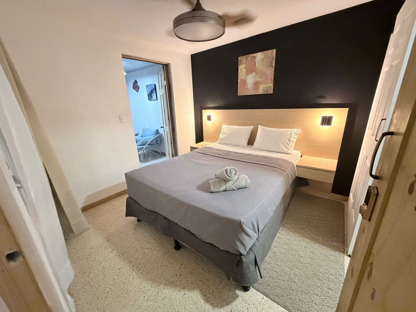 A bedroom with a bed, a door, a wall with a painting, and a ceiling light.