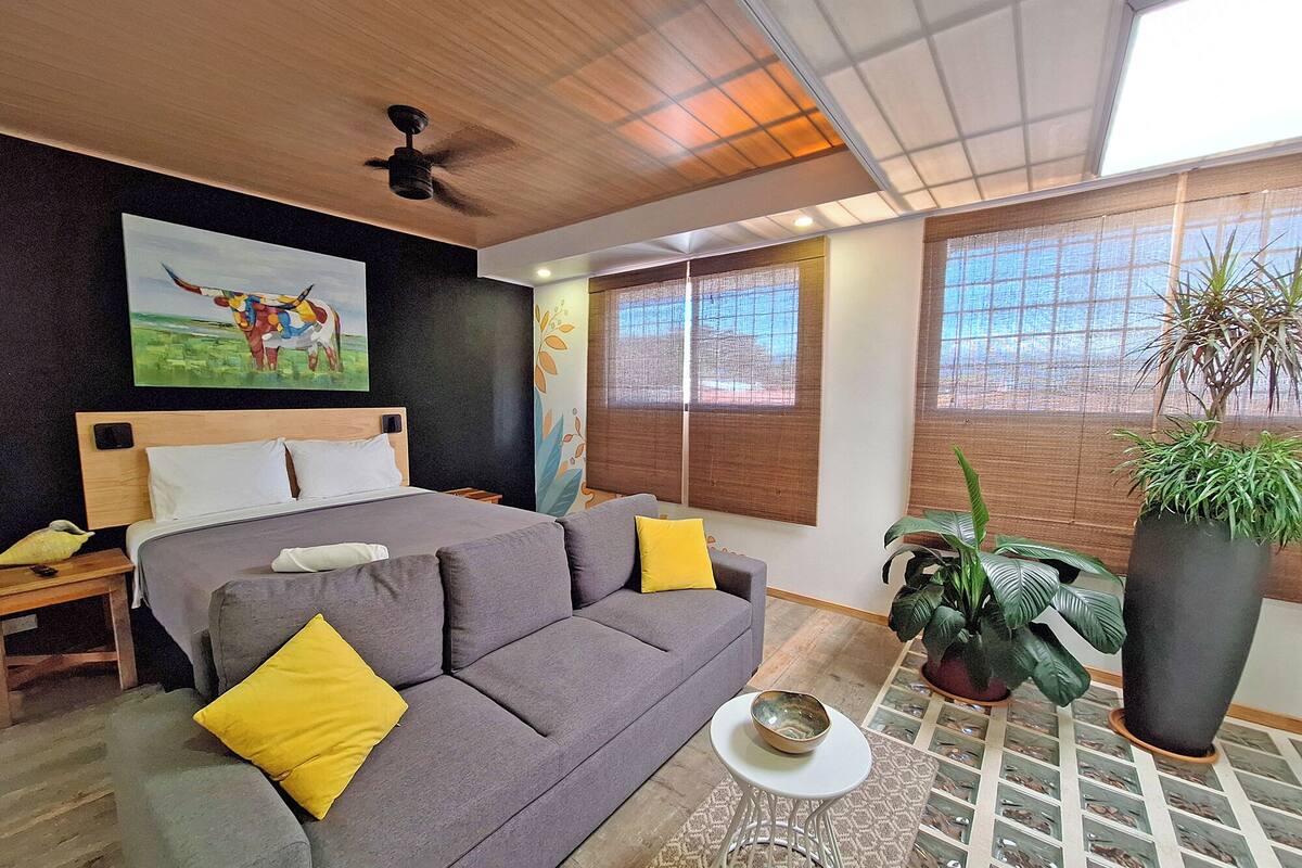 Un dormitorio moderno con una cama, un sofá gris, una mesita blanca pequeña y una planta grande en maceta.