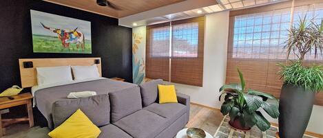 A modern bedroom with a bed, a grey sofa, a small white table, and a large potted plant.