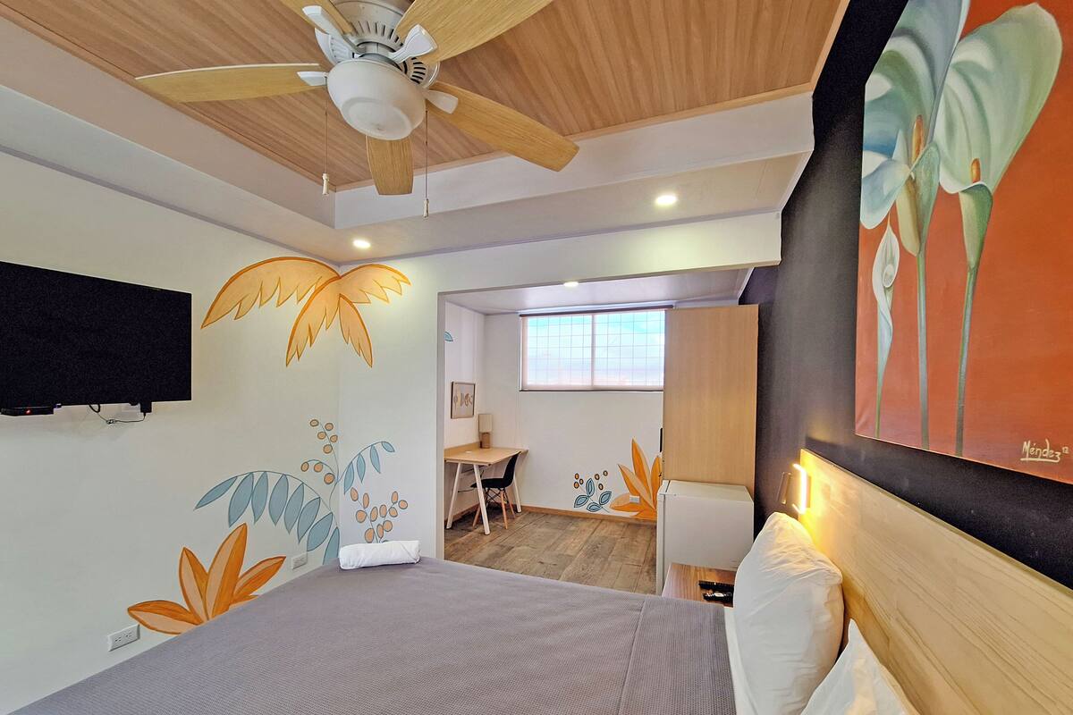 Un dormitorio con cama, ventilador de techo, televisor montado en la pared y un cuadro.