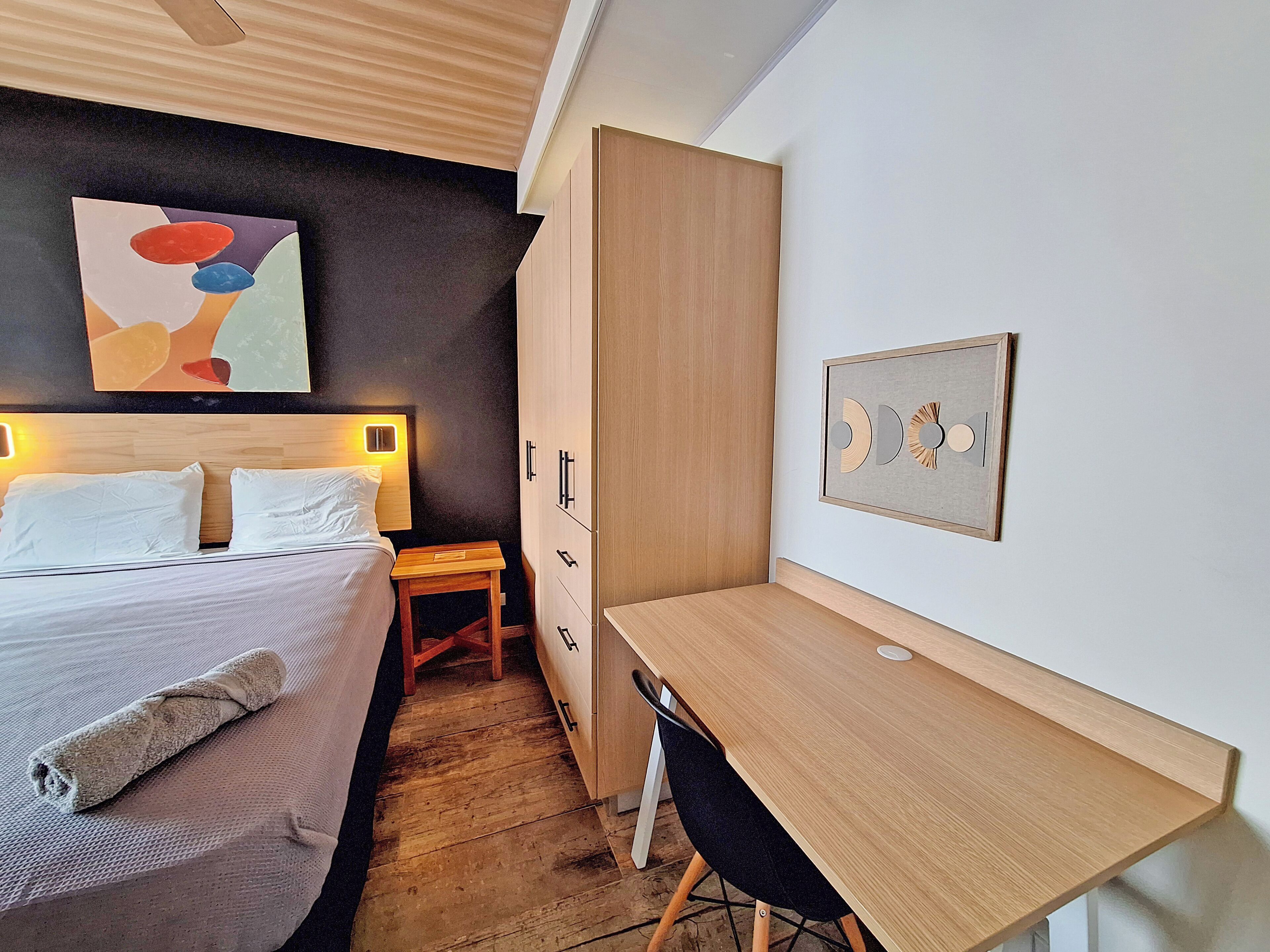 Une chambre à coucher comprenant un lit, une table de chevet et un bureau avec une chaise. On y trouve également une armoire et une œuvre d’art accrochée au mur.